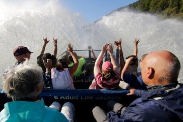 First person view inside the blue jet boat while there is a giant splash in front of them and people with their hands up