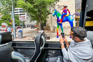 Man taking a photo from the open panoramic tour bus of a rainbow colored sculpture of a cowboy on his horse