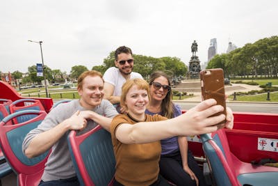 A woman in a yellow shirt takes a selfie of herself and three friends on the top level of a City Sightseeing Philadelphia double decker tour bus.