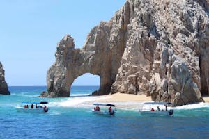 a group of people on a rock next to a body of water with Arch of Cabo San Lucas in the background