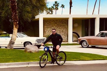 a guest stands in front of a white house in palm springs with a bike on a group tour