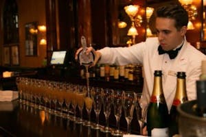 a bar tender pouring cocktails into twenty glasses lined along a bar