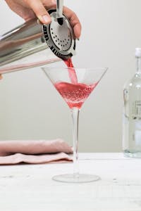 a person holding a cocktail shaker pouring red cocktail into a martini glass