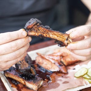 hands that are holding juicy, saucy bbq ribs with pickles on the side