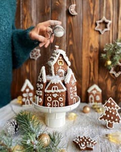 a person sprinkling powdered sugar over a completed gingerbread cottage