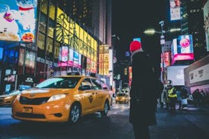 a man walking in a red beanie at night in times square with a taxi driving by