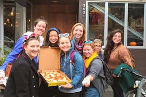 A group of women holding up a pizza box open with a pizza in it.