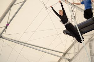 An adult female hangs on a flying trapeze at an indoor gym. The woman is an amateur trapeze artist.