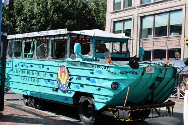 A bright blue Amphicoach bus that is labeled the "Boston Duck Tours Land and Water". The name of the bus is Annie Aquarium. These tour buses can drive on land and water.
