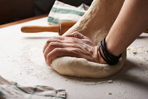 a close up of a person's hands kneading dough and a rolling pin in the back ground