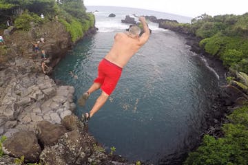 An older gentleman in swim trunks jumping off a cliff into a pool of water in Hana