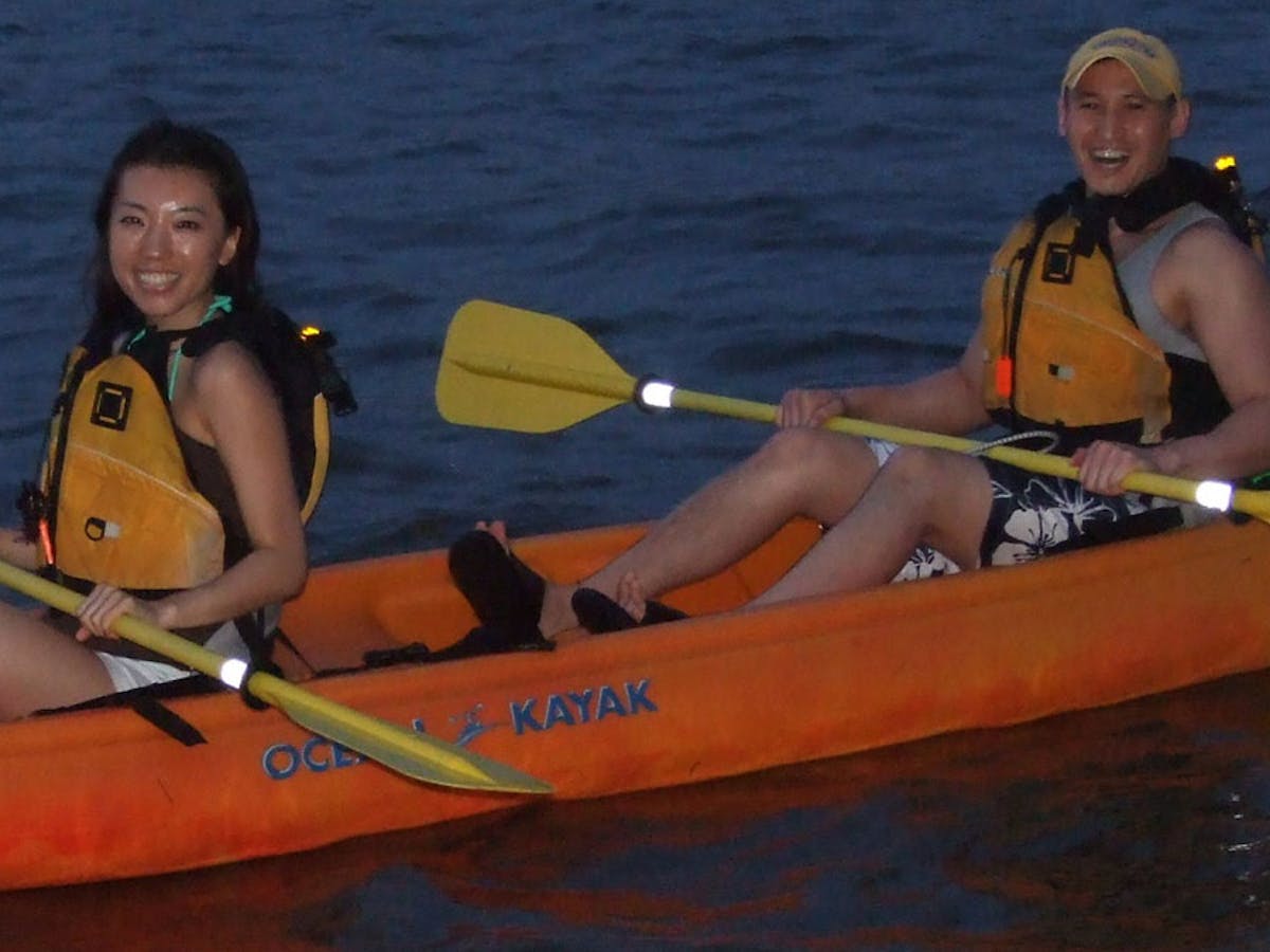 A couple in an orange kayak with yellow paddles smiling and kayaking at night