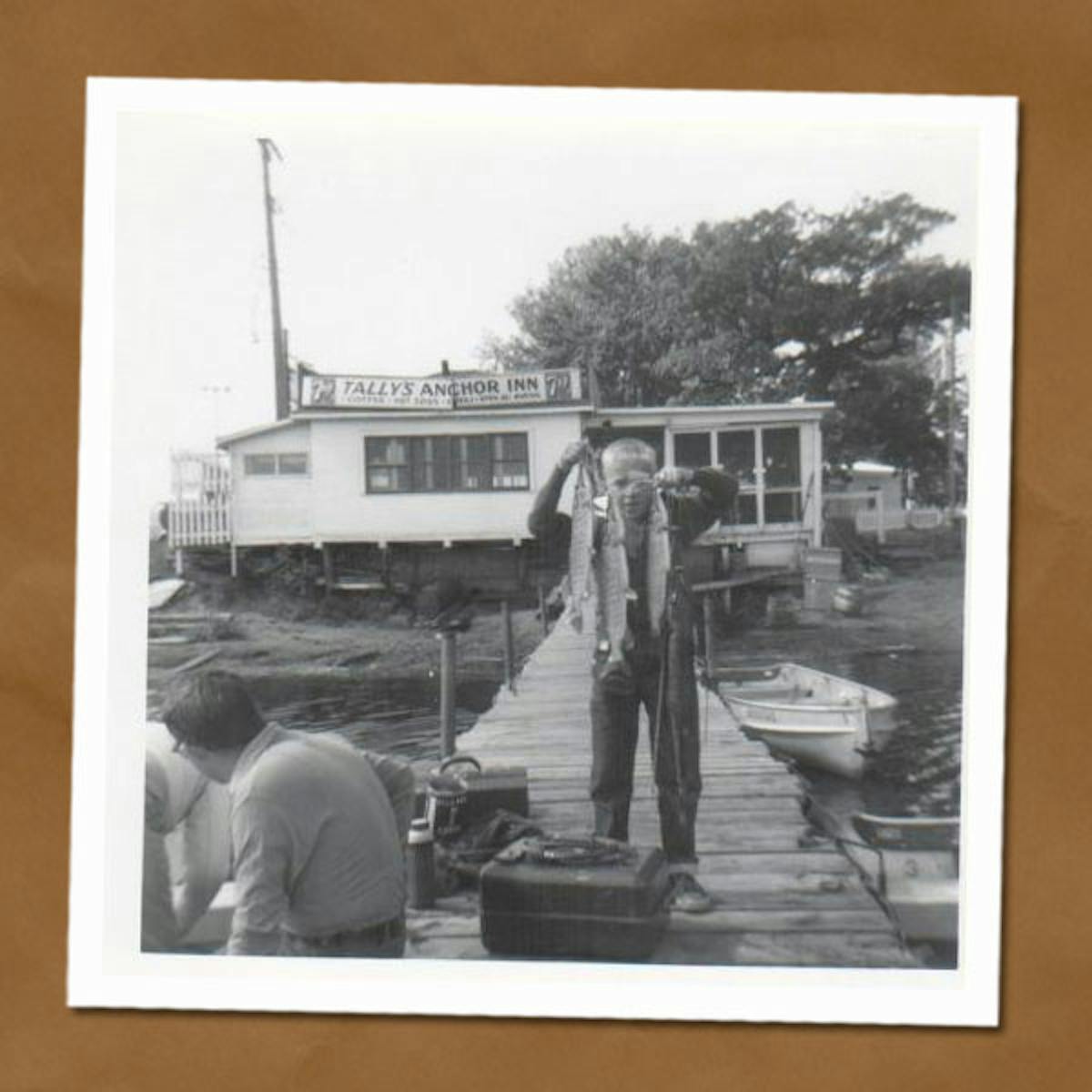 Black and white photo of man standing on dock holding up string of fish