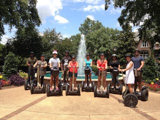 Guests smiling on Segways in front of a beautiful water fountain. Green lush trees are surrounding the water fountain.