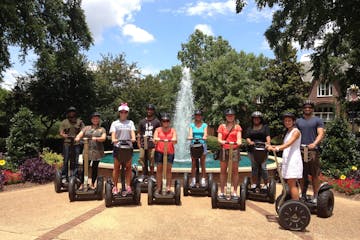 Guests smiling on Segways in front of a beautiful water fountain. Green lush trees are surrounding the water fountain.