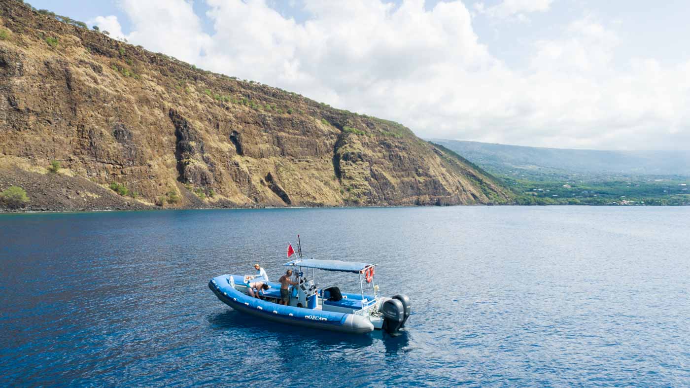 a snorkel boat with a few passengers sits on calm water off a dramatic tall rocky cliff