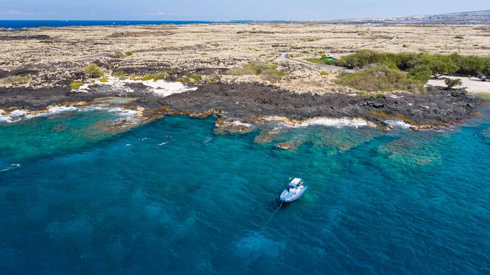 aerial view of the kona coast with reef visible through the water and snorkelers near the rugged lava coastline. A boat floats in the water nearby