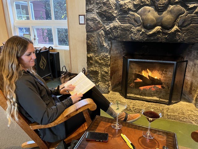 a woman sitting in a rocking chair in warm clothing holding a book. A lava rock fireplace in the background and a pile of wood. A table sits next to the woman with martinis on it.