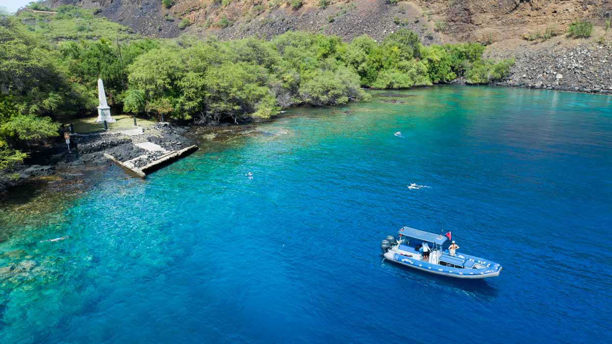 a boat sits in the calm ocean water just off shore from a white obelisk nestled among some trees with a cliff behind