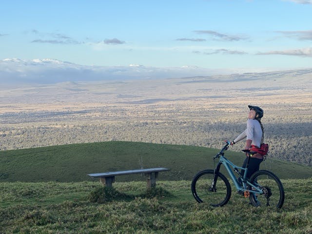 woman on a mountain bike posing awkwardly on a grassy hill with yellow dead grassy view in the background and clouds on mountains behind