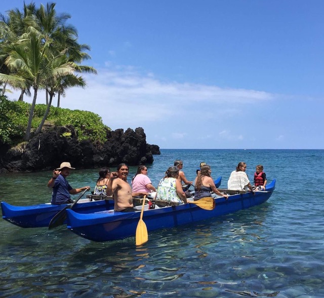 a double hull canoe sit in the water with people holding paddles inside and a rocky coast with a palm tree in the background