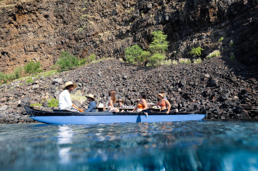 people sitting in a double hulled canoe in the ocean in front of a steep cliff