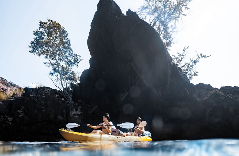 a man and woman sitting in an open top kayak in the ocean with a large pointy rock behind
