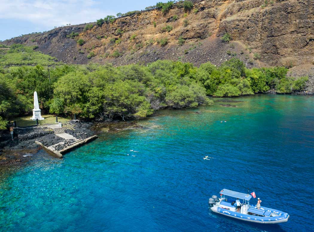 an aerial view of the captain cook monument with a boat floating in the ocean just offshore and some snorkelers in the water nearby