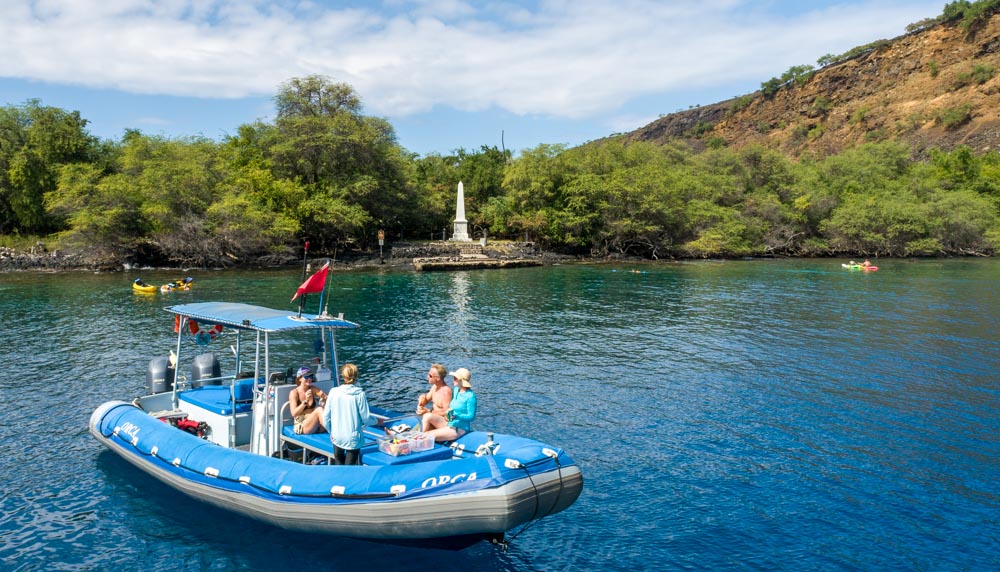 people sit on a small boat in the ocean talking. There are kayaks in the water behind and a whilte obelisk of the captain cook monument on shore