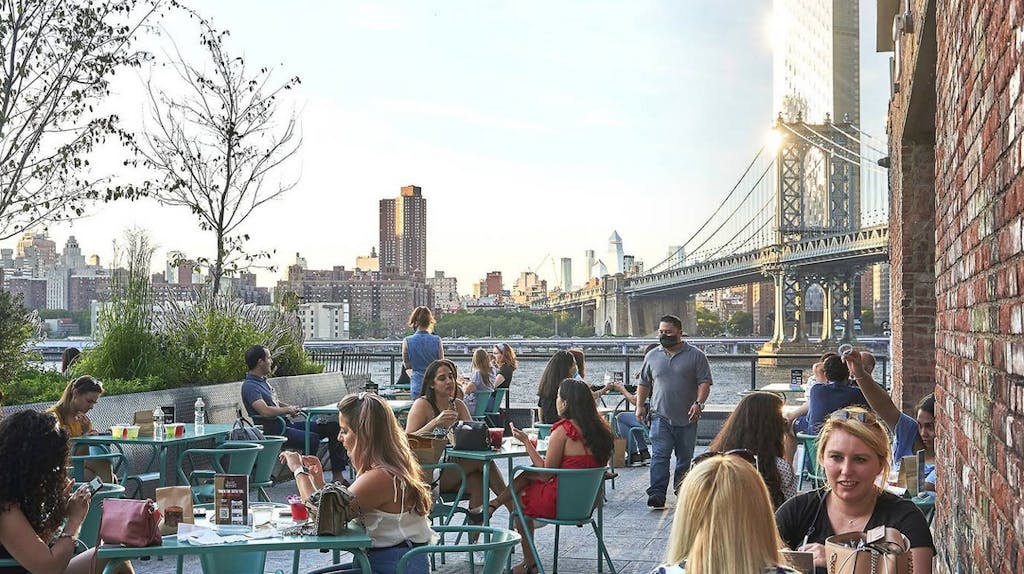 people sitting outside at tables and eating with the manhattan bridge in the view