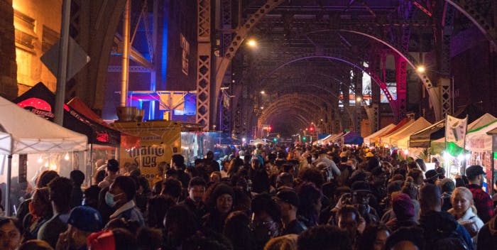 large crowd walking the street at night visiting food vendors under a bridge