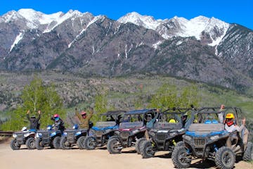 A group of RZR's and ATV's with people on them are parked in front of big mountains in the background