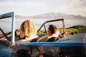 two men in a car with a surfboard looking at the ocean with mountains in the background