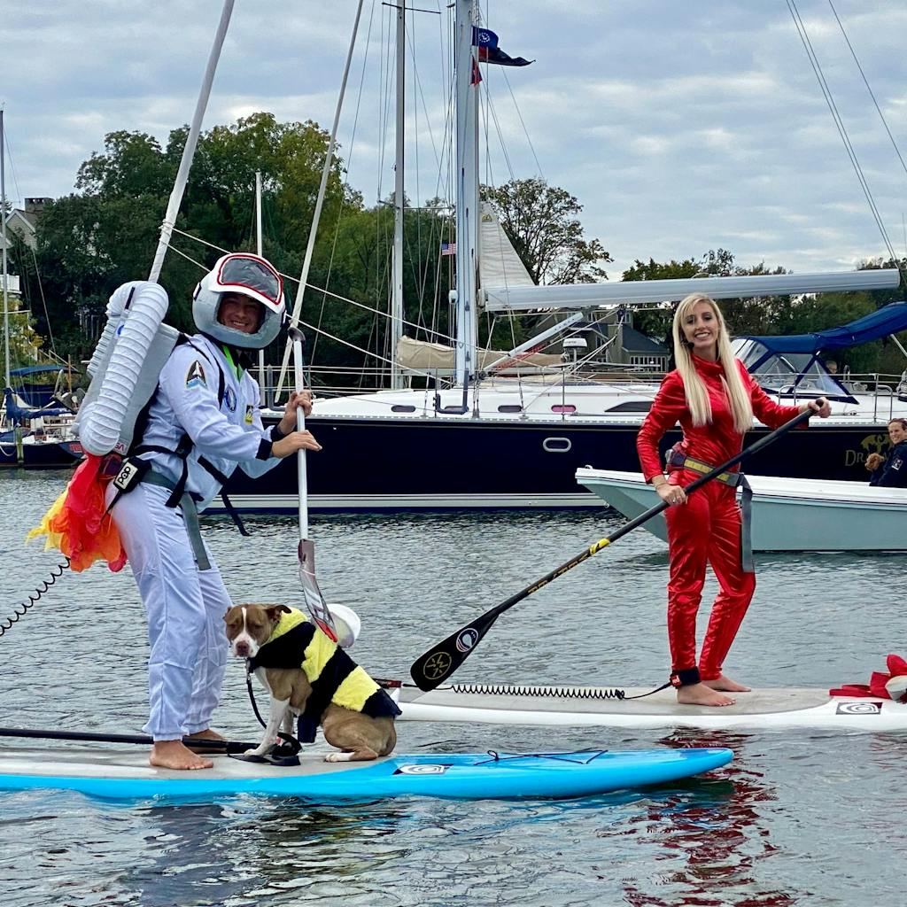 A man dressed as an astronaut on a paddle board with a dog, next to a woman dressed as Britney Spears on a paddle board on the water.