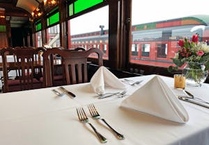 A Dining Table Is Set Nicely With A Folded Napkin And Flowers On The Dining Train Car.
