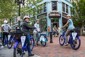 A group of people are standing over their electric bikes smiling at the camera while on a food tour in a city. They are in front of a restaurant called the Chocolate Lounge on a brick paver sidewalk.