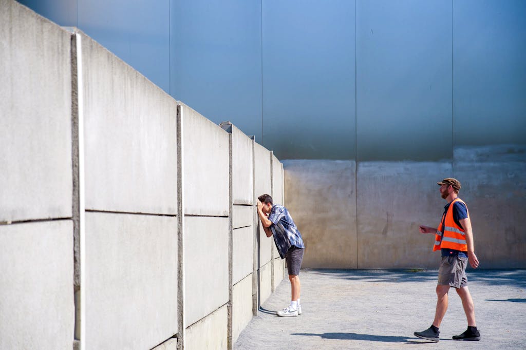 Eine Person schaut durch die Mauer an der Mauergedenkstätte Bernauer Straße, dahinter sieht man einen Tourguide mit einer orangenen Weste