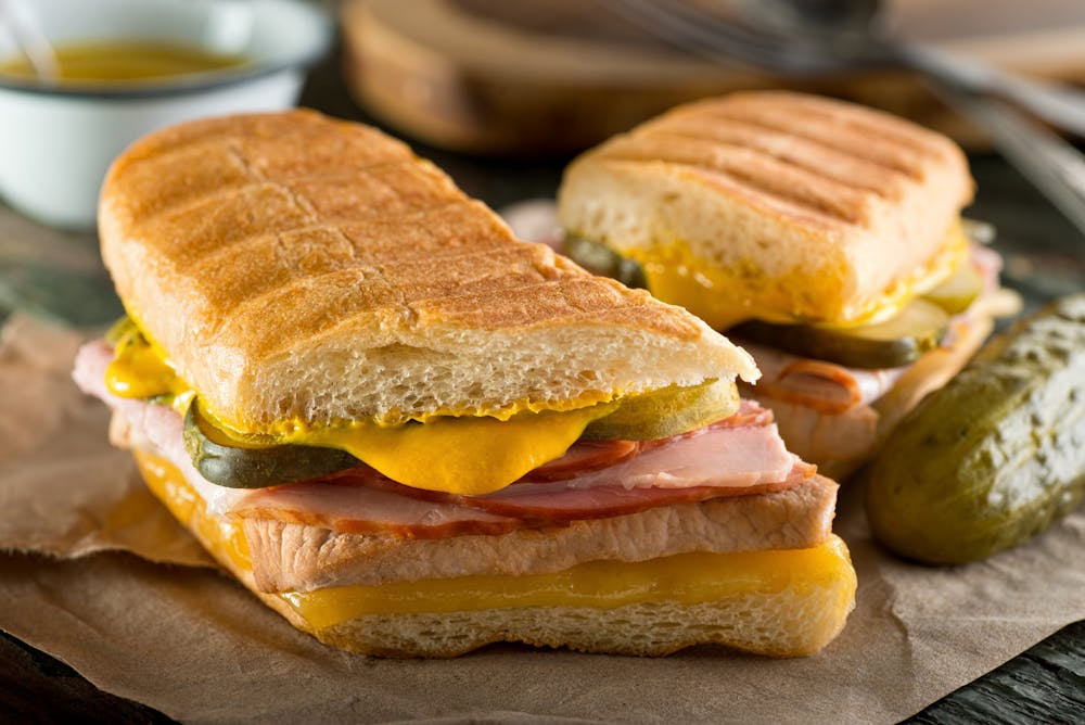 An authentic cuban sandwich on pressed medianoche bread with pork, ham, cheese, pickle, and mustard.