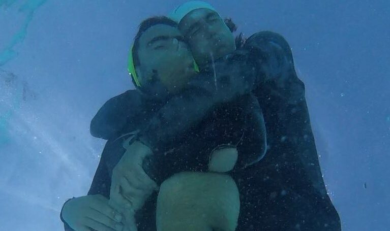 Two divers holding their breath and grabbing onto each other, swimming to the surface
