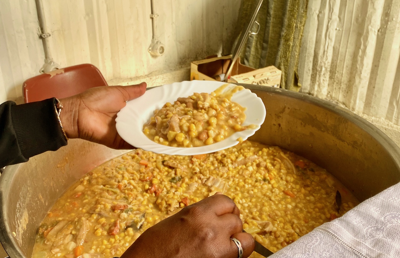 a woman serving a plate of cachupa from a huge pan 