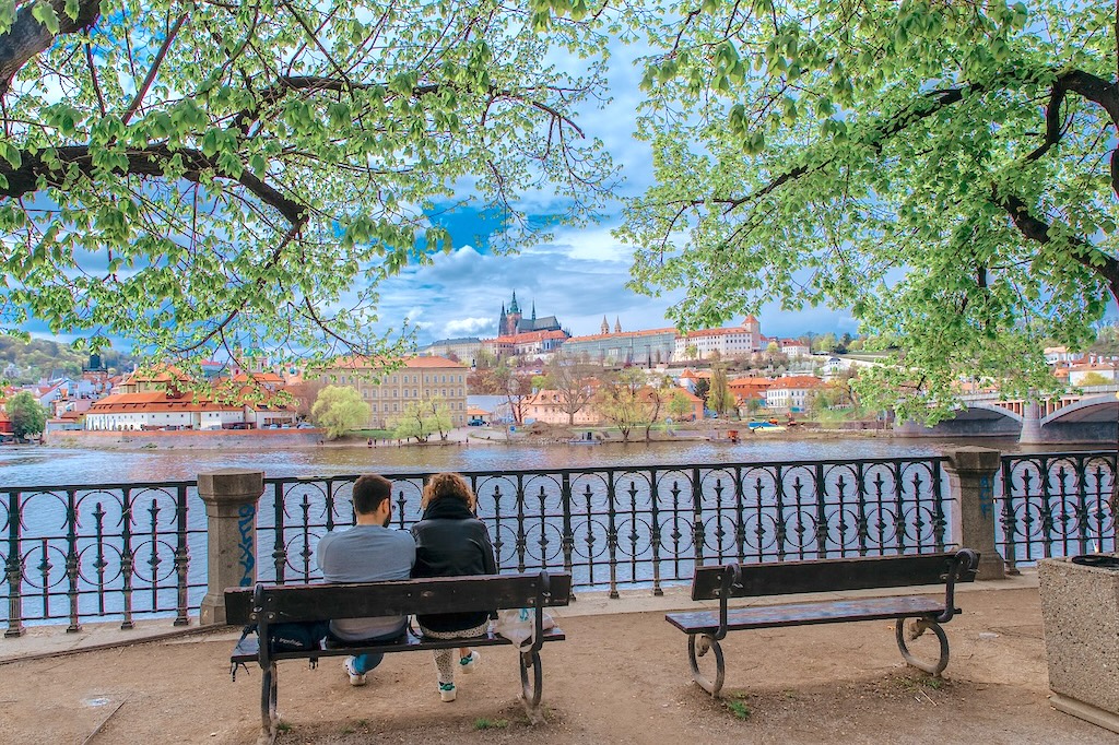 a young couple in love sitting on a bench on the Vltava River bank with a view of the Prague Castle in the background