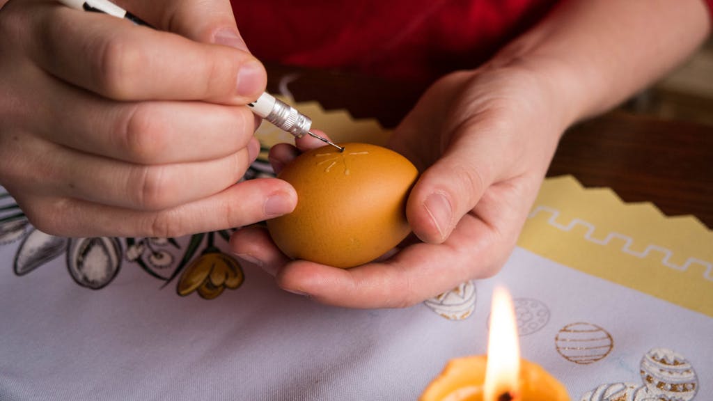 one hand holding a yellow egg and the other decorating it with wax