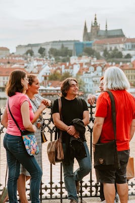A group of tourists facing a tour guide, standing on the Vltava River Bank with a beautiful view of Prague Castle in the background