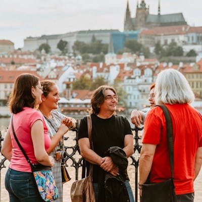 A group of tourists facing a tour guide, standing on the Vltava River Bank with a beautiful view of Prague Castle in the background