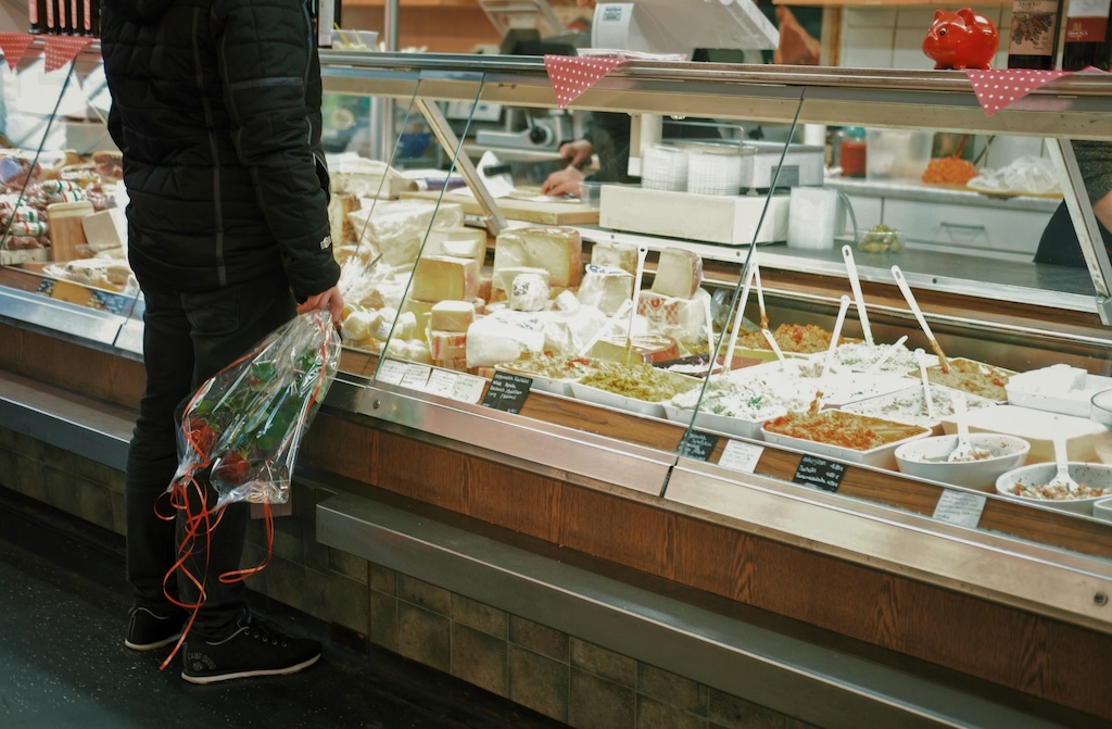 a person holding a bouquet of flowers standing in front of the counter in a classical Czech deli store, looking at their variety of cold salads