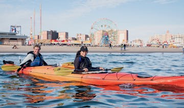 a smiling man and woman sitting in an orange double sea kayak in front of Coney Island Beach
