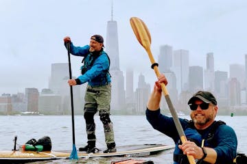 a man riding a kayak and a man riding a SUP with the Freedom Tower in the background