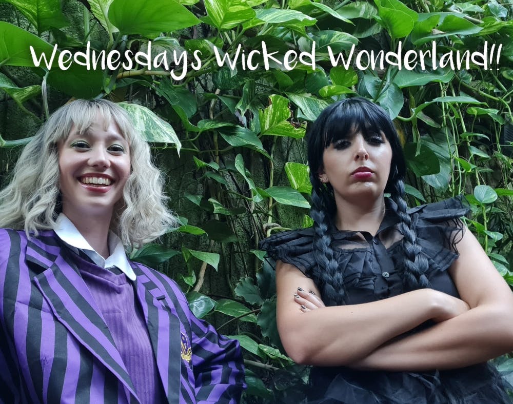 Two girls dressed as Enid and Wednesday pose in front of a green plant wall