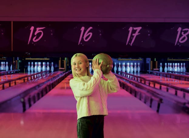 a girl holding a bowling ball in the air, in front of bowling lanes