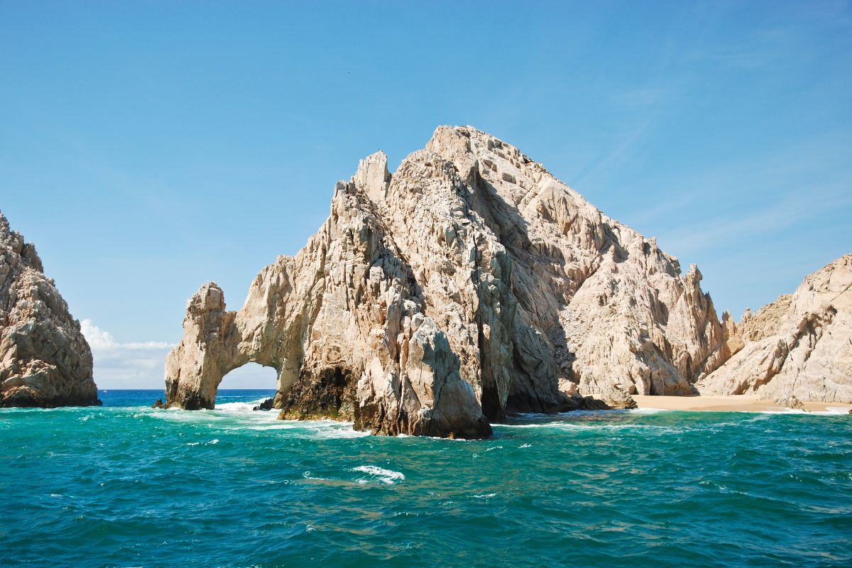 a close up of a rock near the ocean with Arch of Cabo San Lucas in the background